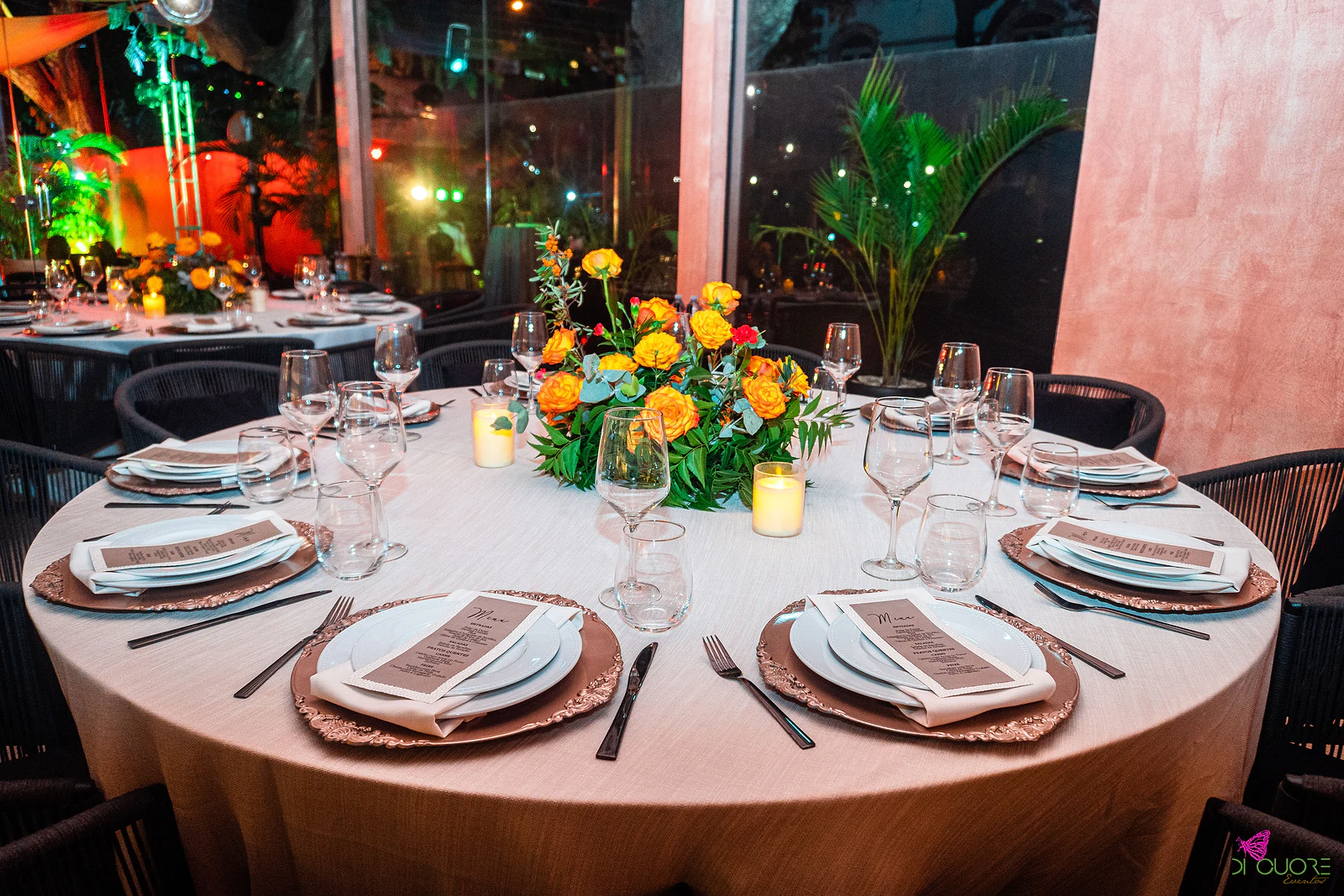 Mesa redonda de toalha clara posta com sousplats cobre e menus, com centro de flores laranja e amarelas, junto a uma parede cor-de-rosa e a uma palmeira