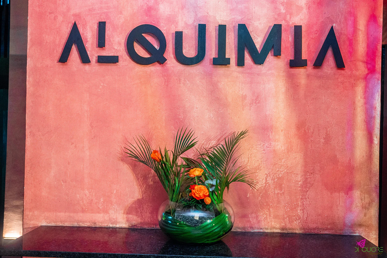 Letreiro “ALQUIMIA” em letras pretas sobre uma parede cor-de-rosa texturada, com uma taça de vidro redonda com palmas e rosas laranja sobre um tampo escuro