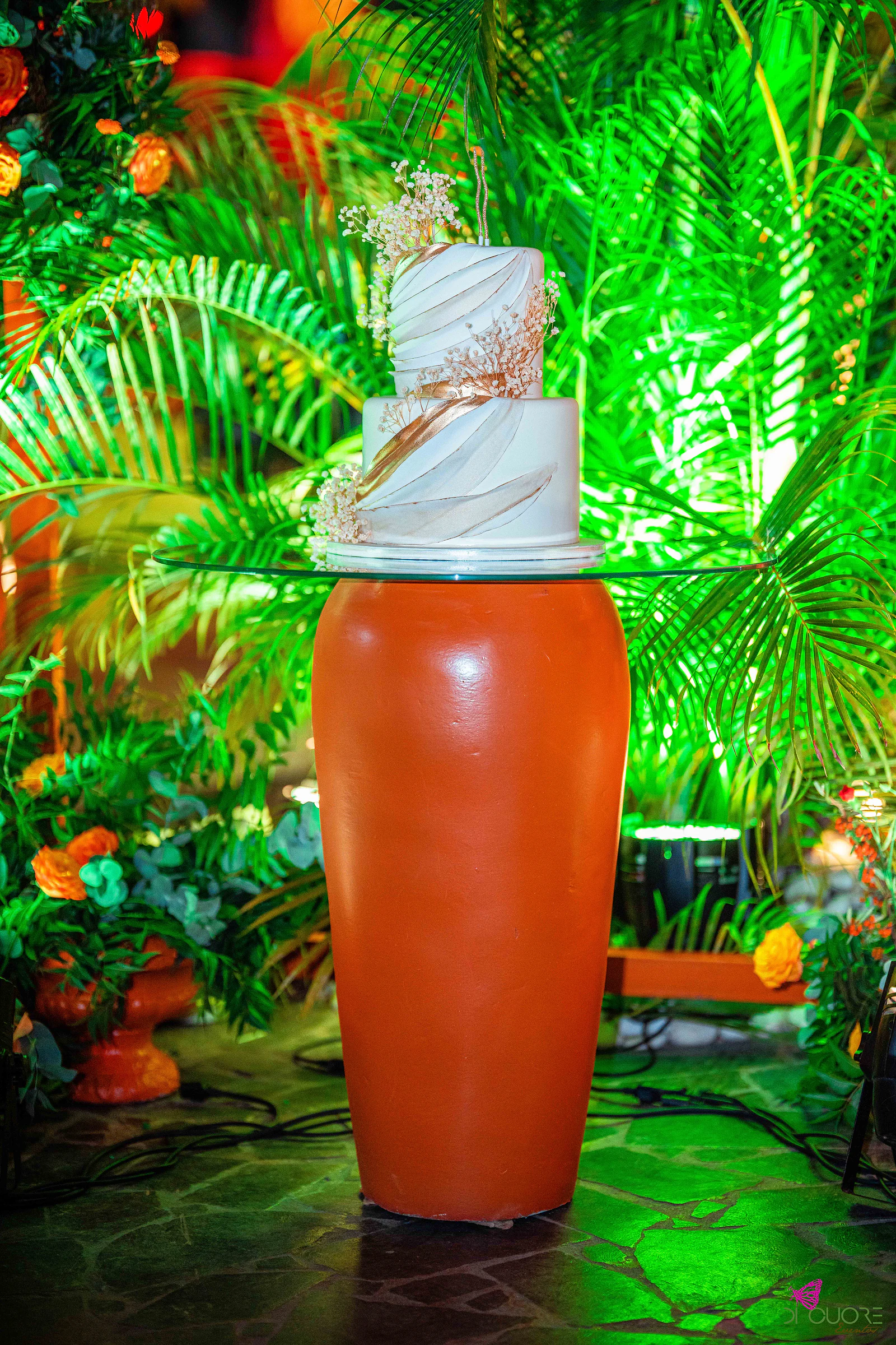 Bolo de duas camadas com ondas esculpidas e pormenores cobre, sobre uma jarra laranja, com palmeiras iluminadas a verde ao fundo