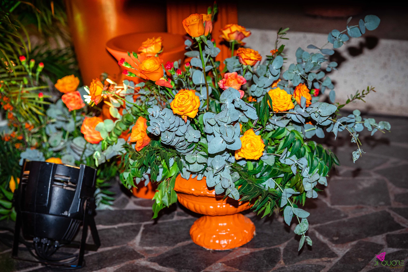 Arranjo de rosas laranja e amarelas com eucalipto numa urna laranja sobre pavimento de pedra, com um projector preto ao lado