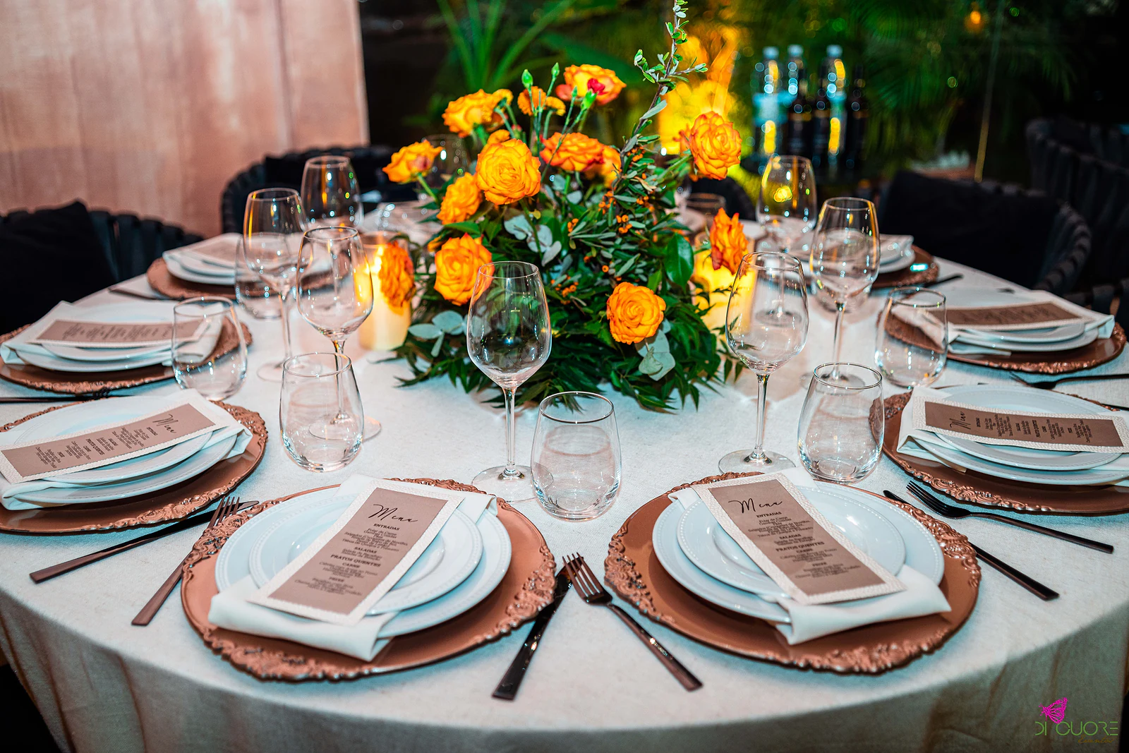 Pormenor da mesa posta, com sousplats cobre, menus impressos, copos e centro de rosas laranja com eucalipto