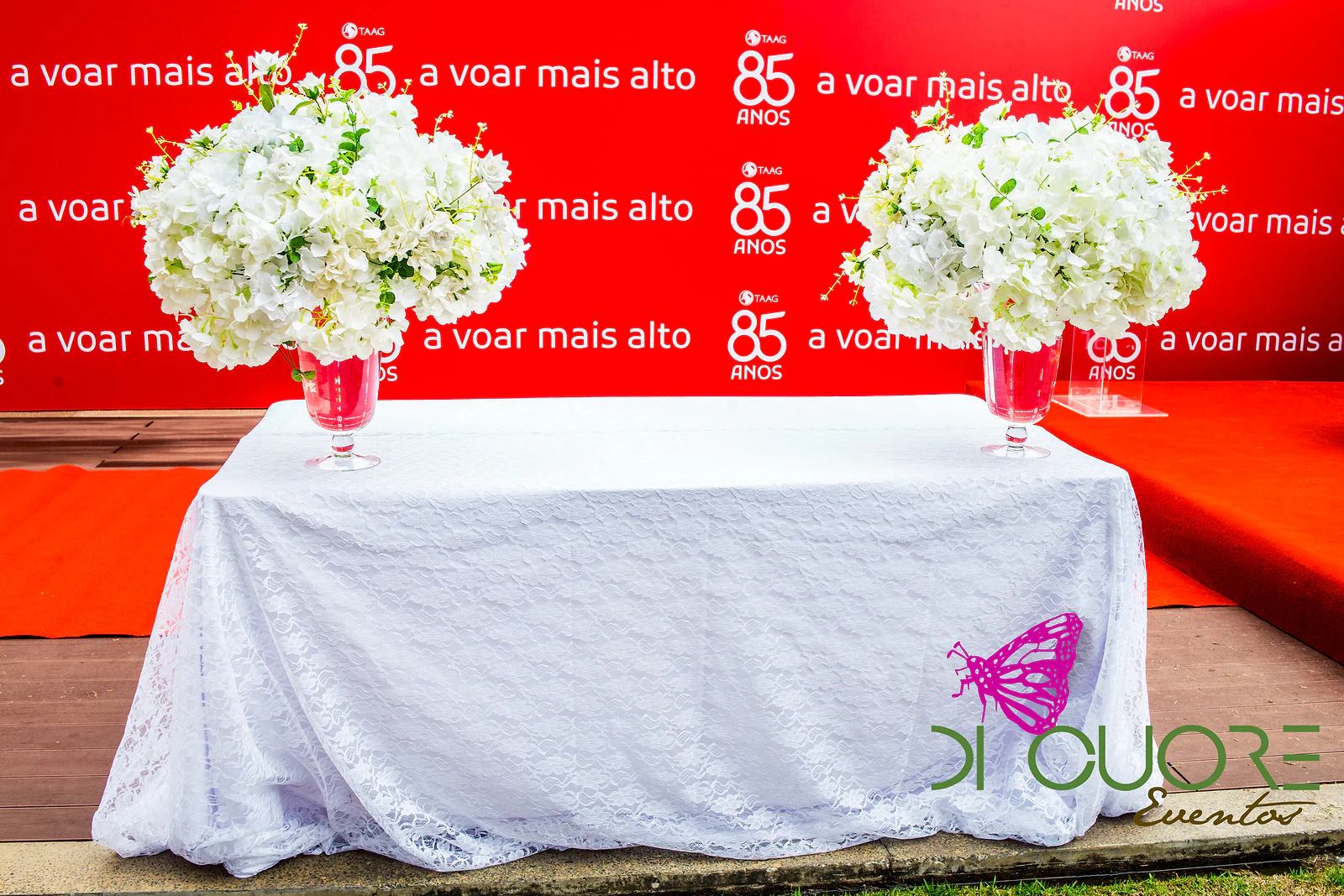 Mesa de toalha de renda branca com dois grandes ramos de flores brancas em vasos de pé alto, diante da parede vermelha com o lema “a voar mais alto”