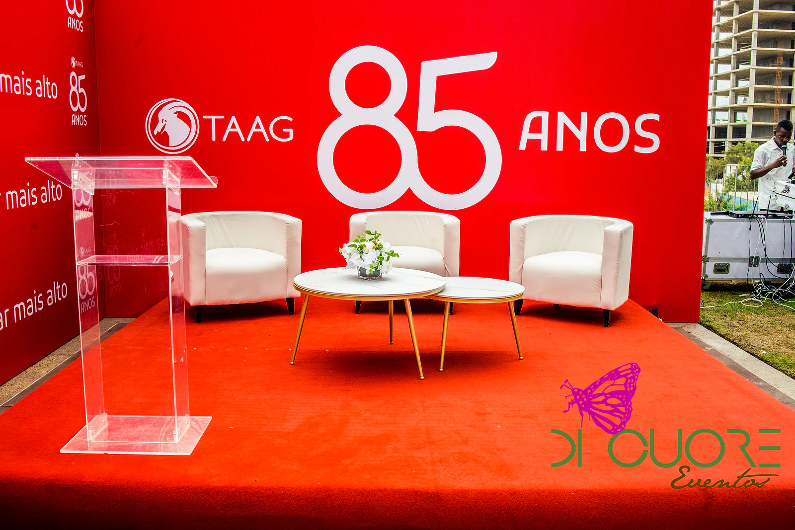 Red-carpeted stage with three white armchairs, coffee tables on gold legs, a small flower arrangement and an acrylic lectern, in front of the 85 anos wall