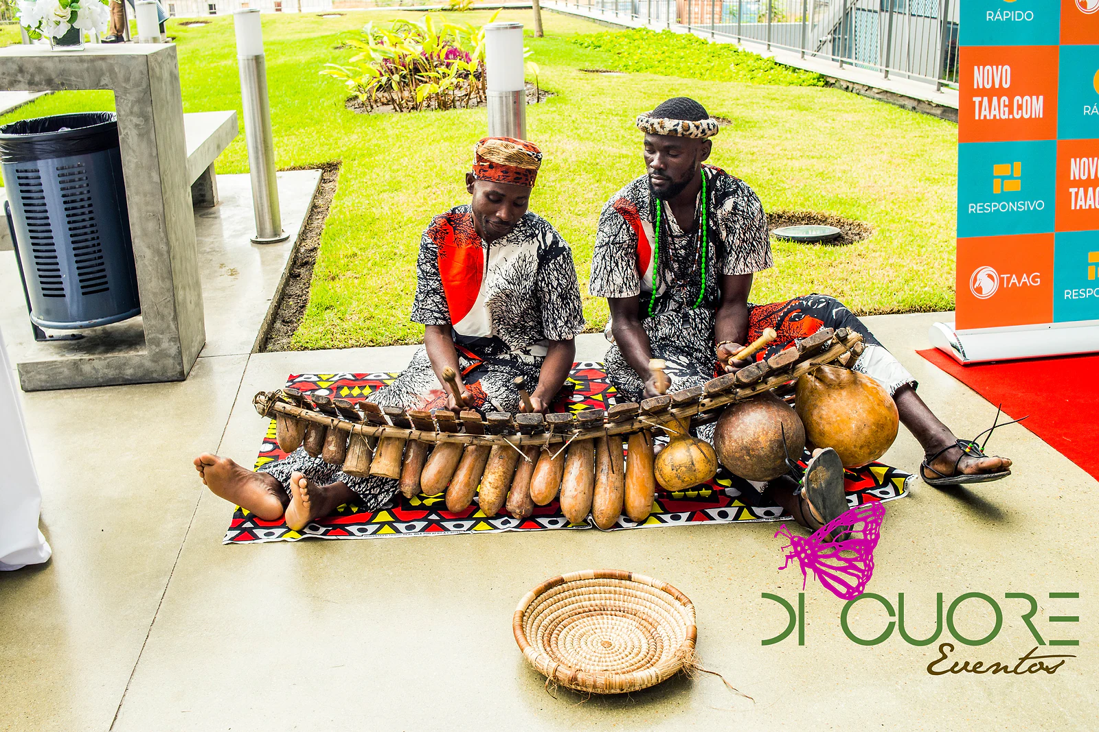 Dois músicos de traje de padrão africano a tocar marimba de cabaças sobre um pano colorido, com um cesto de palha no chão e relvado ao fundo