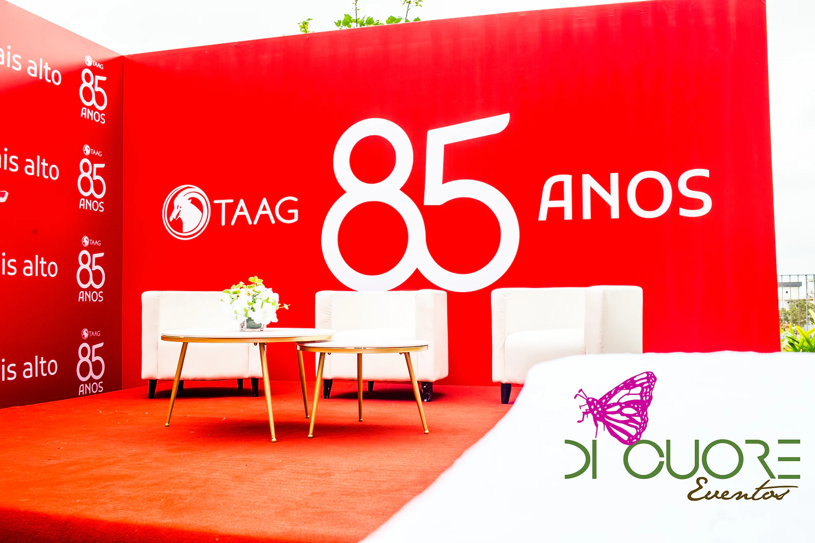 Poltronas brancas e mesas de centro douradas com um ramo de flores, sob o logótipo TAAG e o 85 anos em branco sobre fundo vermelho