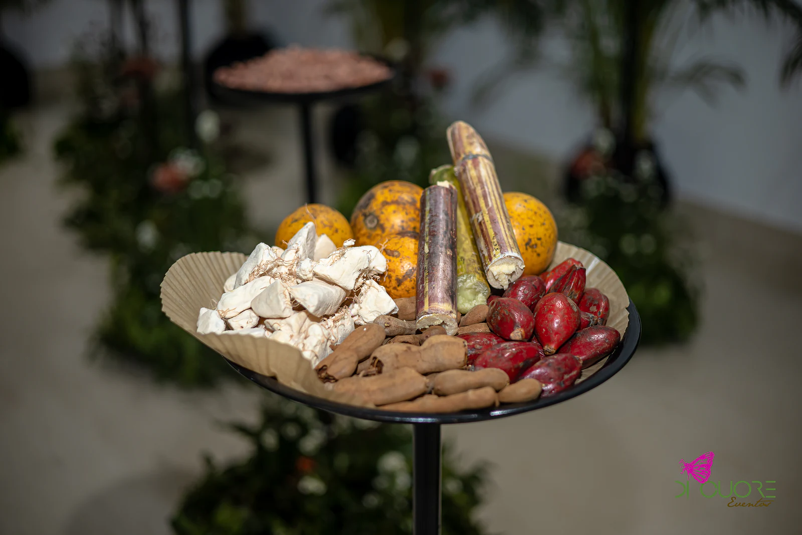 Prato num pedestal preto com cana-de-açúcar, frutos cor de laranja e vermelhos e vagens, entre folhagem desfocada