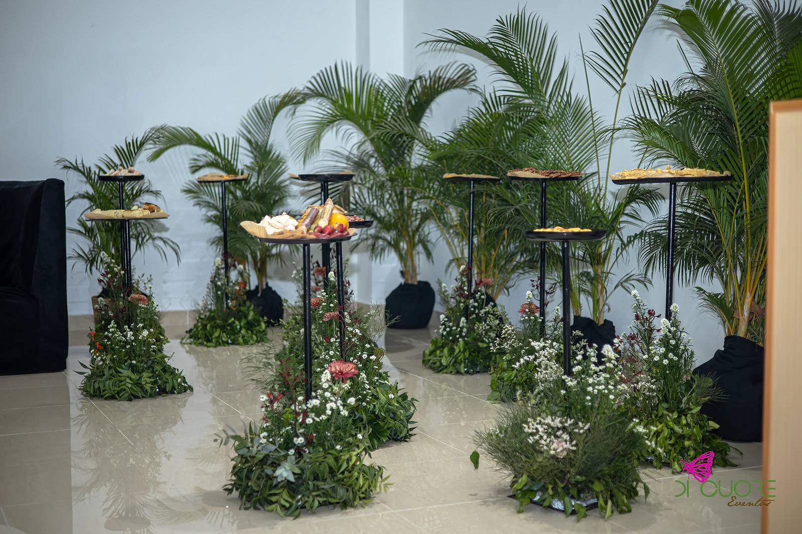 Ilhas de folhagem e flores brancas aos pés de pedestais pretos com petiscos, entre palmeiras