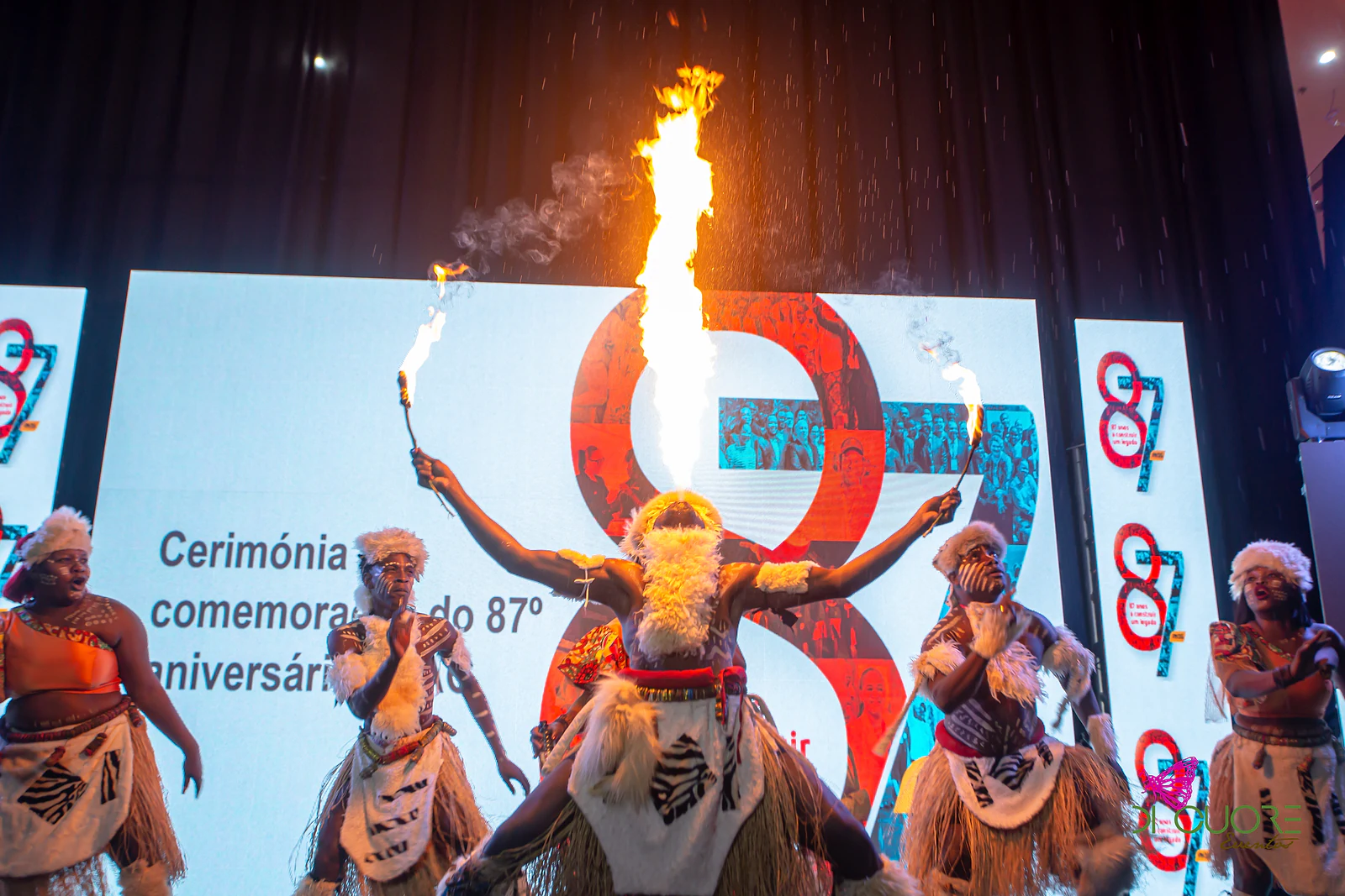 Artista a cuspir uma labareda alta durante uma actuação de dança tradicional, com outros bailarinos de trajes típicos ao lado e o número 87 num ecrã ao fundo