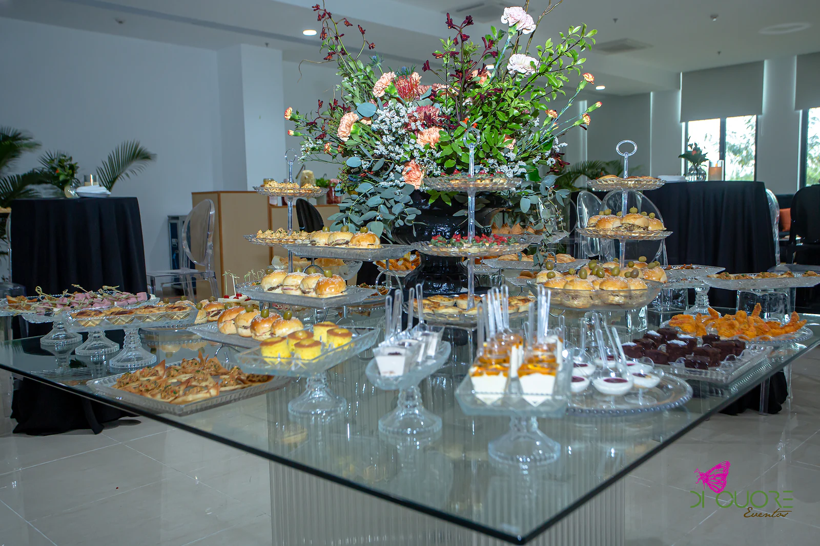 Mesa de vidro com doces e salgados em bases de vidro e, ao centro, um grande arranjo de flores cor de laranja e brancas