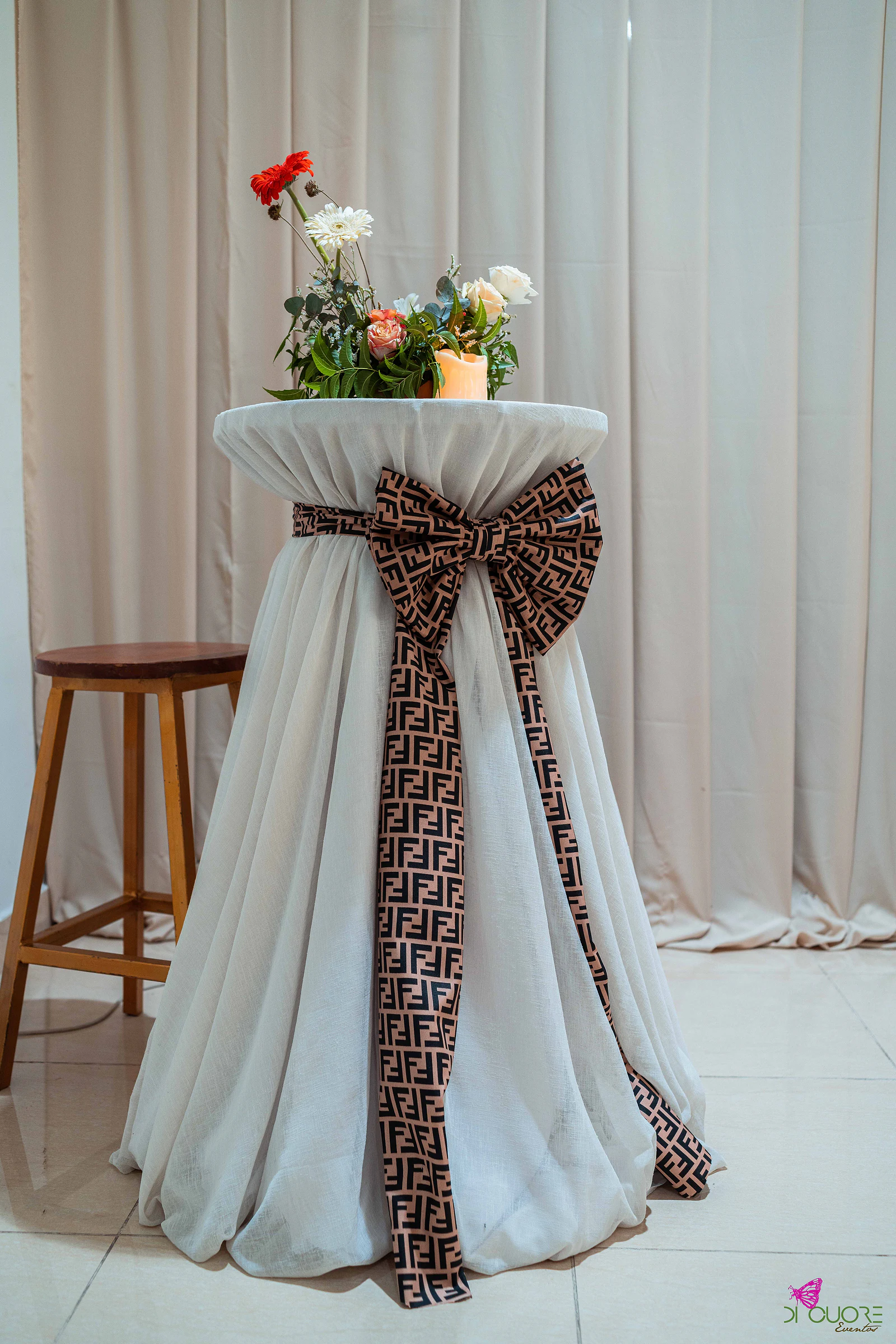 Mesa alta com saia branca, grande laço castanho de padrão geométrico, ramo de flores com vela e banco de madeira