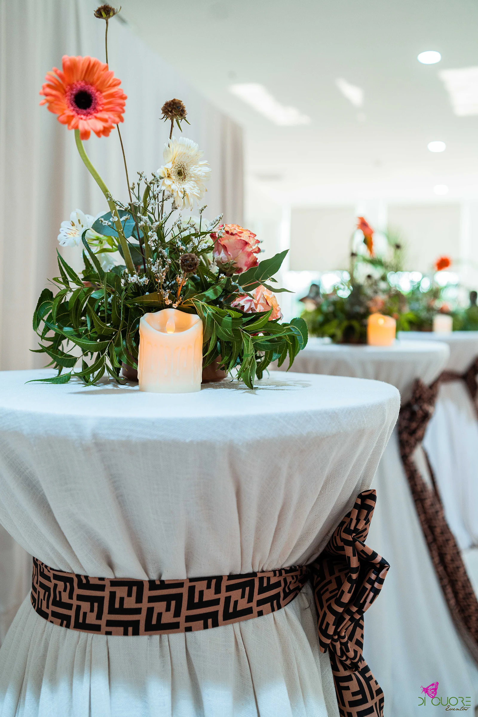 Mesa alta com saia branca cintada por um laço castanho, ramo de gerbera, rosas e folhagem com uma vela, e mais mesas iguais ao fundo