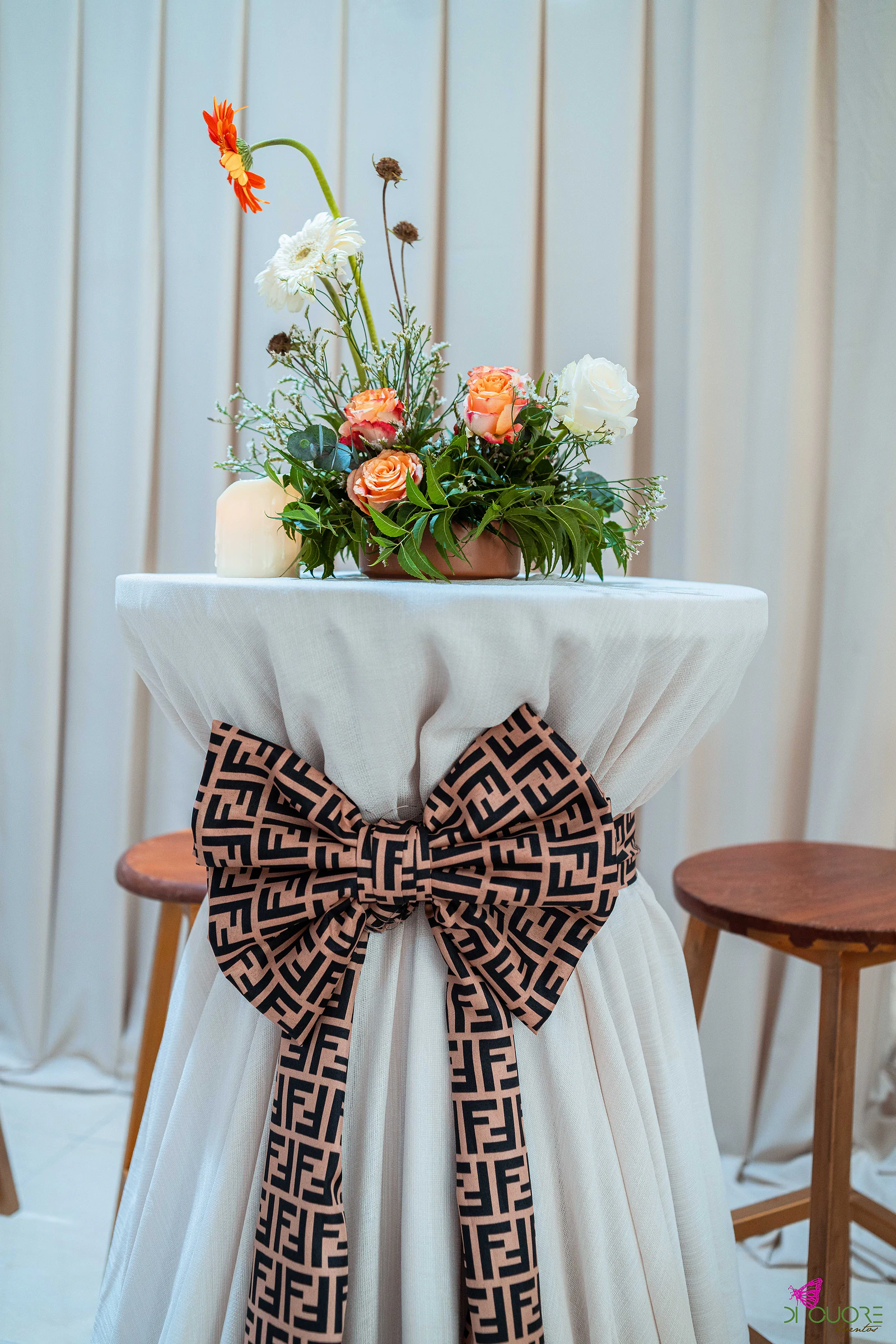 Mesa alta com saia branca e grande laço castanho de padrão geométrico, ramo de rosas cor de pêssego, rosas brancas e gerbera, e bancos de madeira dos lados, diante de uma cortina clara