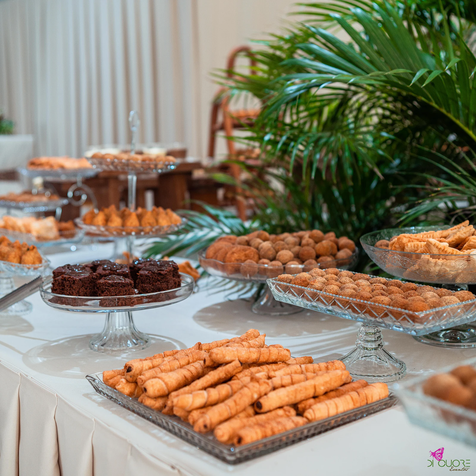 Bandeja de vidro com salgados enrolados, pratos com bolinhos e brownies de chocolate em bases de vidro, sobre toalha branca, com palmeiras ao fundo