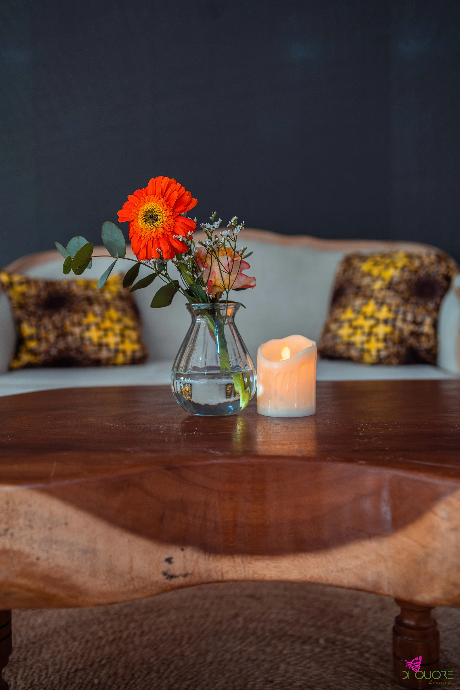 Pormenor da mesa de centro em madeira de bordas naturais, com uma gerbera laranja num jarro de vidro e uma vela, diante do sofá e de almofadas amarelas e castanhas