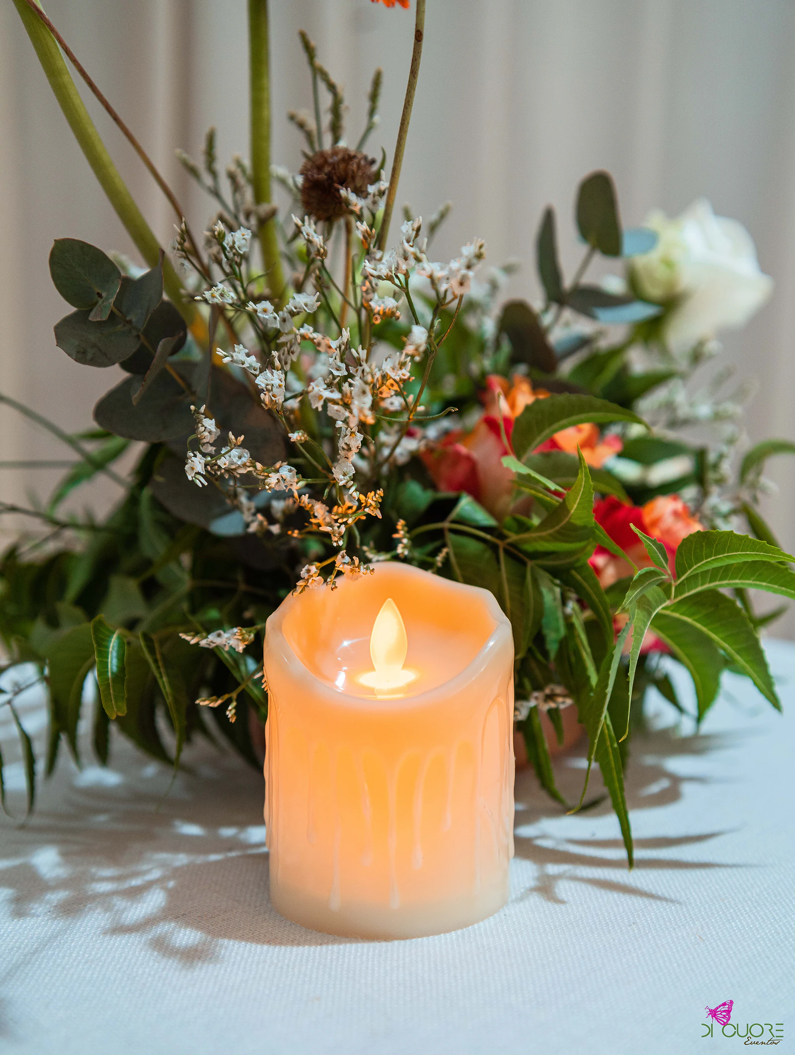 Vela de luz quente junto a um ramo de flores laranja e folhagem, sobre toalha branca