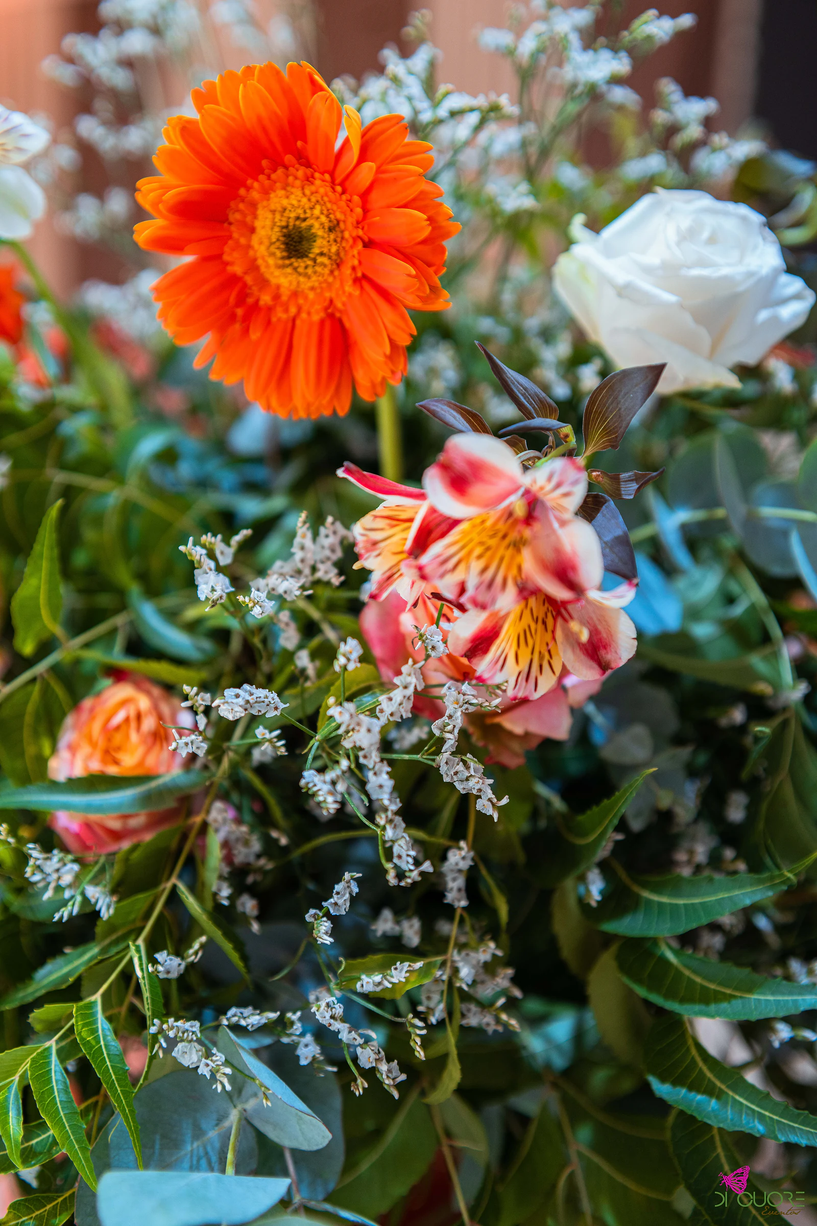 Pormenor de uma gerbera laranja, alstroemérias e uma rosa branca entre a folhagem