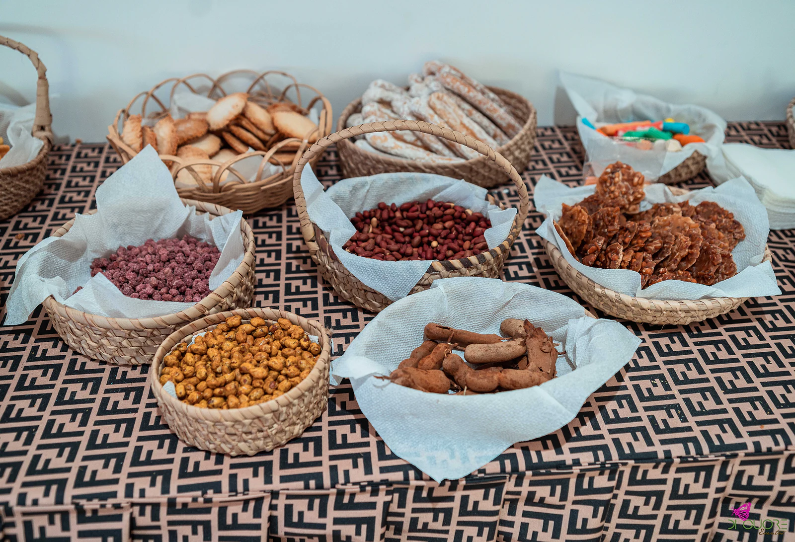 Cestos de vime forrados a papel branco com biscoitos, frutos secos, tamarindo e doces, sobre o tecido castanho estampado