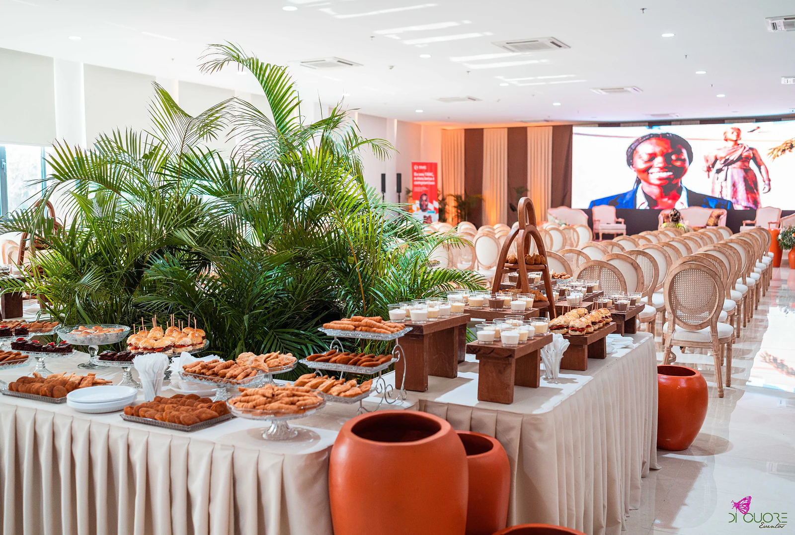 Mesa de buffet com saia branca entre palmeiras, bancos de madeira com sobremesas e grandes vasos de barro, com o salão ao fundo