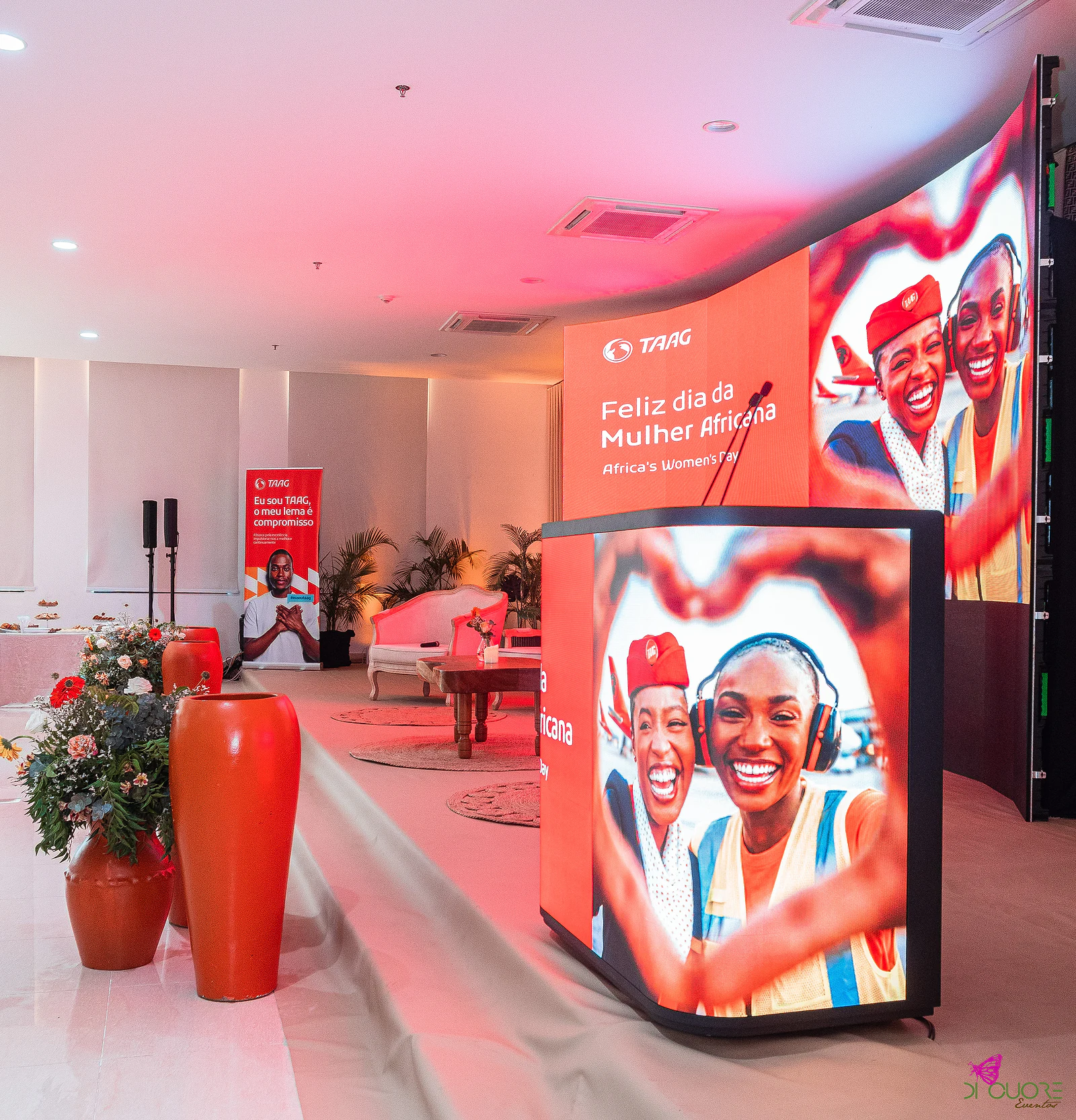 Púlpito com ecrã a mostrar duas mulheres sorridentes, junto a um painel laranja com a mensagem «Feliz dia da Mulher Africana», numa sala iluminada em tons rosados, com vasos de barro cor de laranja em primeiro plano