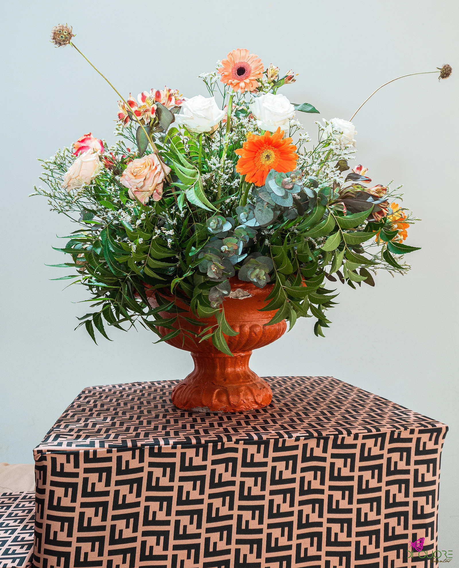 Taça de barro cor de laranja com gerberas, rosas e eucalipto, sobre um cubo forrado a tecido castanho estampado