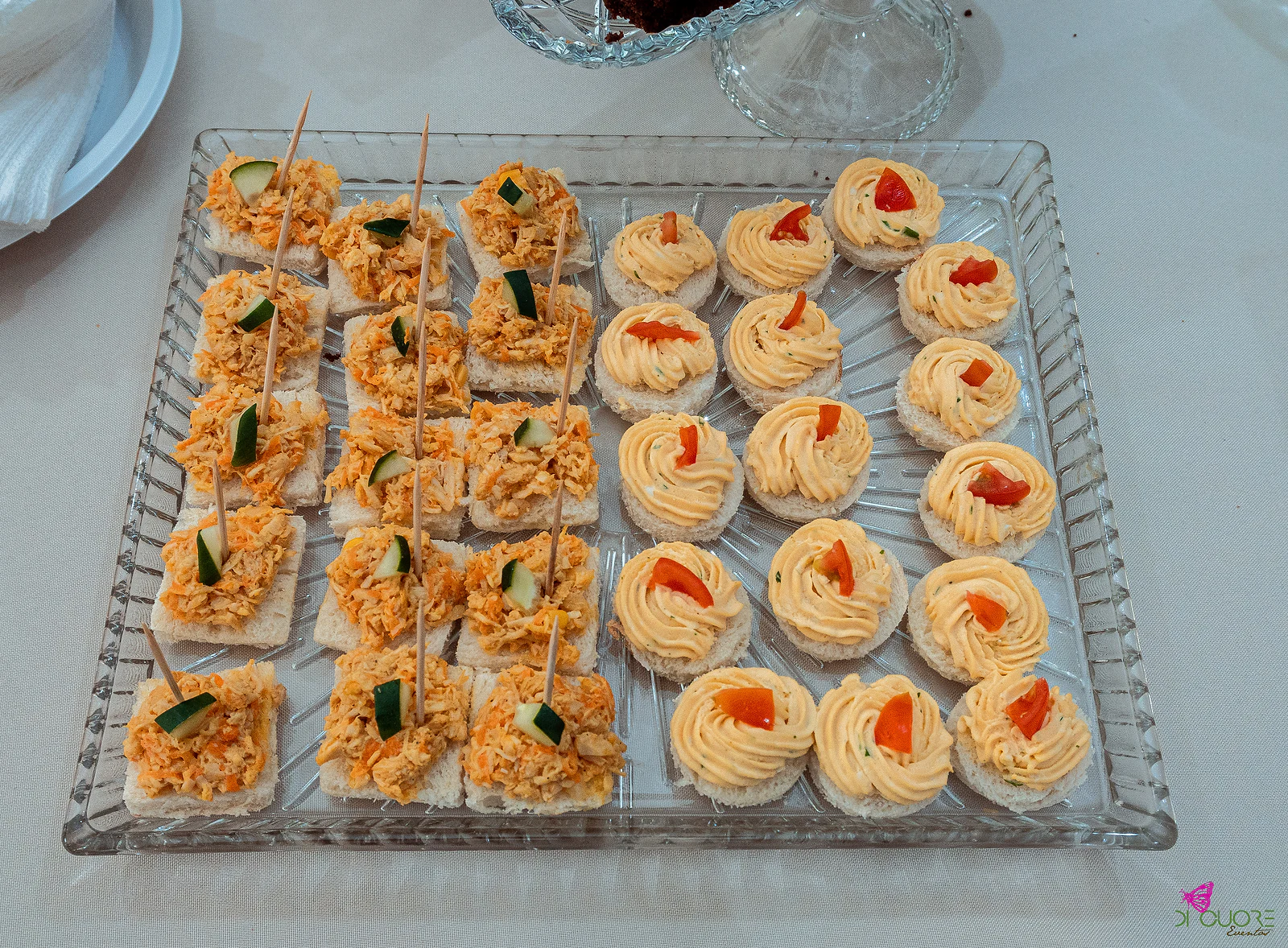 Bandeja de vidro com canapés de pão de forma, uns com pasta de frango e pepino, outros com creme alaranjado e tomate