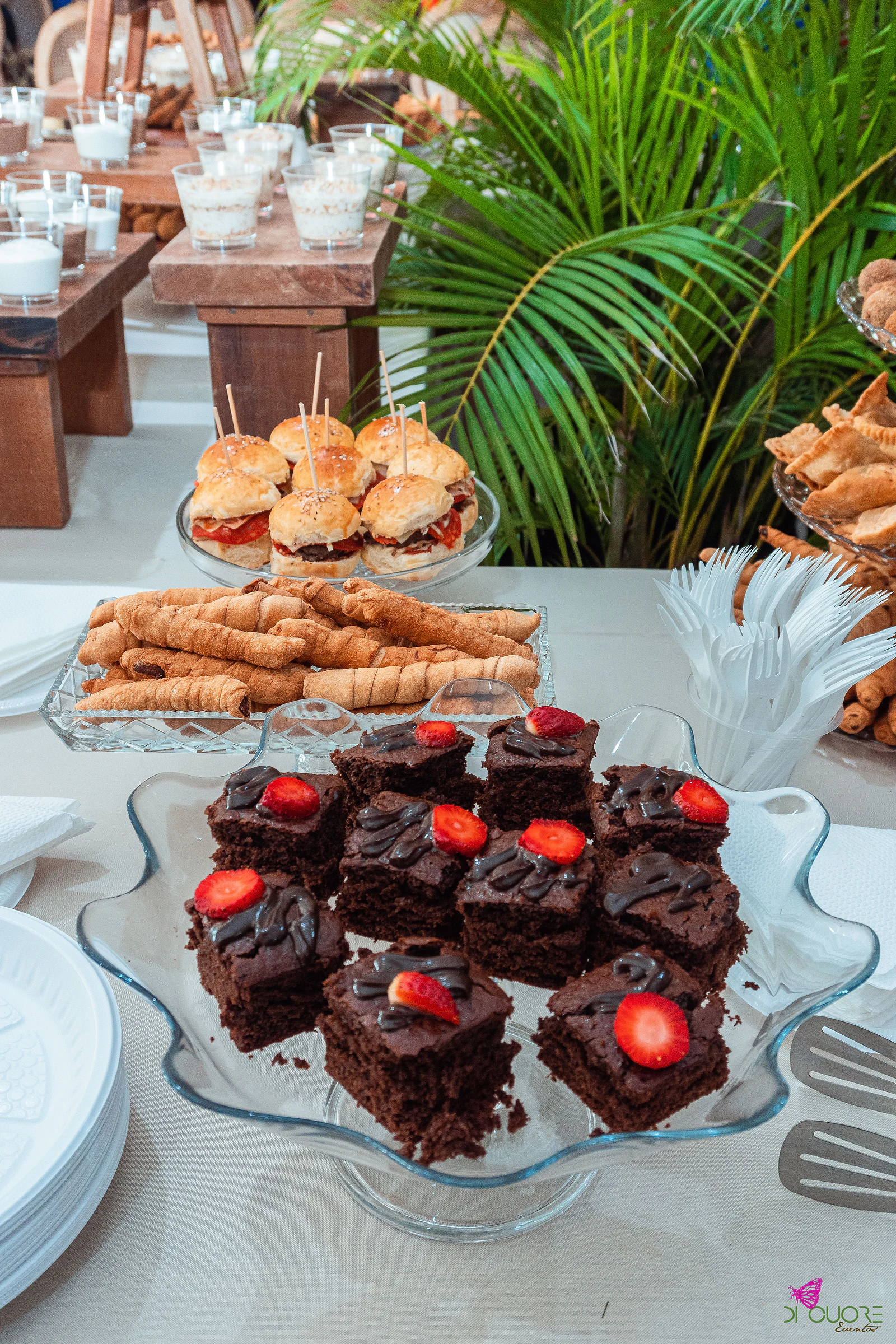 Taça de vidro com quadrados de bolo de chocolate e morango, atrás um prato de salgados enrolados e mini-hambúrgueres com palitos, com palmeiras e bancos de madeira ao fundo