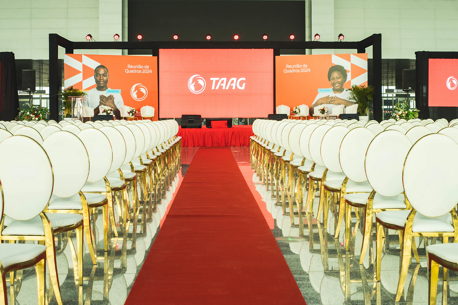 Red carpet between rows of white chairs with gold legs, leading to the stage with TAAG’s orange and red screens