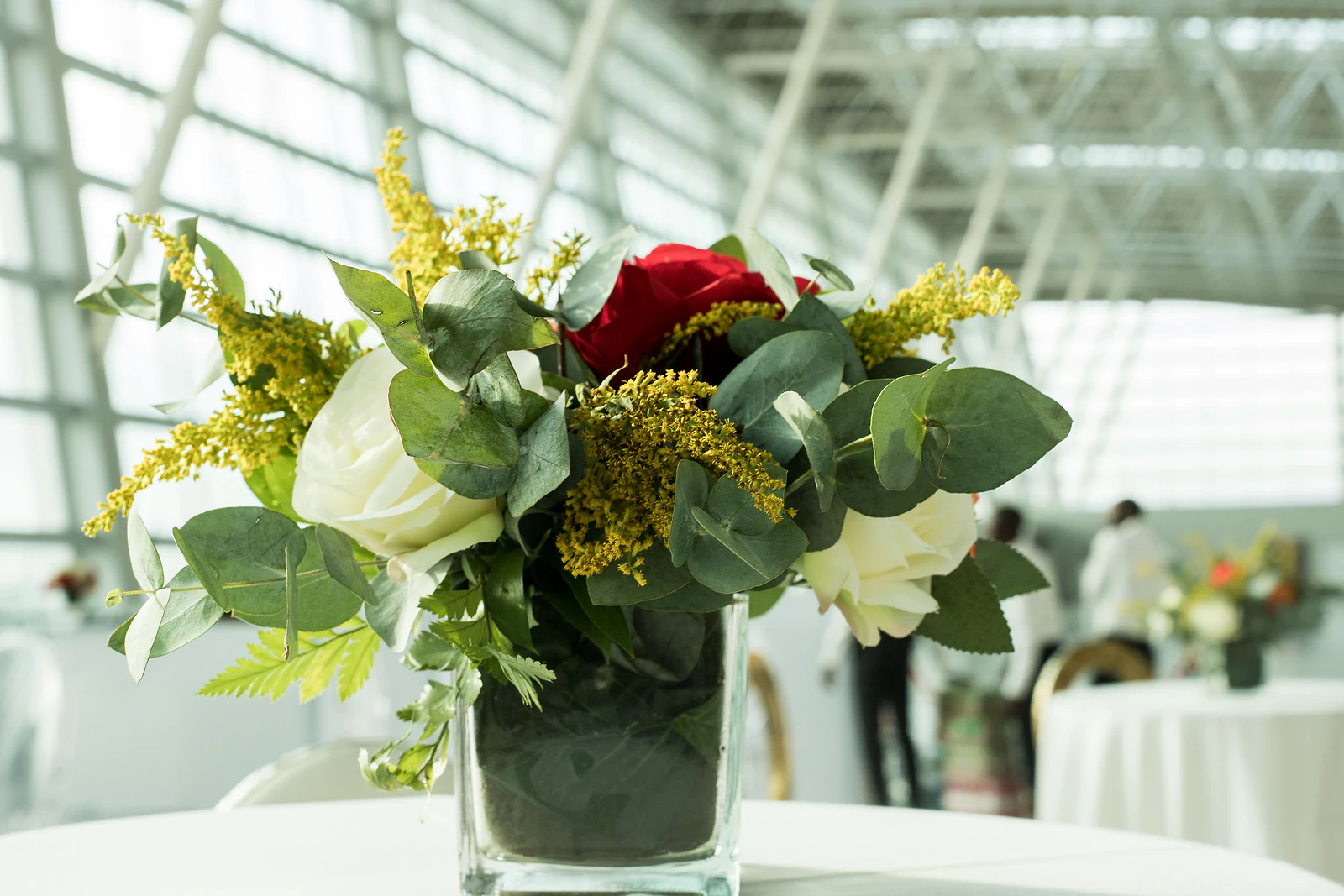 Small arrangement of a red rose, white roses, solidago and eucalyptus in a square glass vase on a white-clothed table