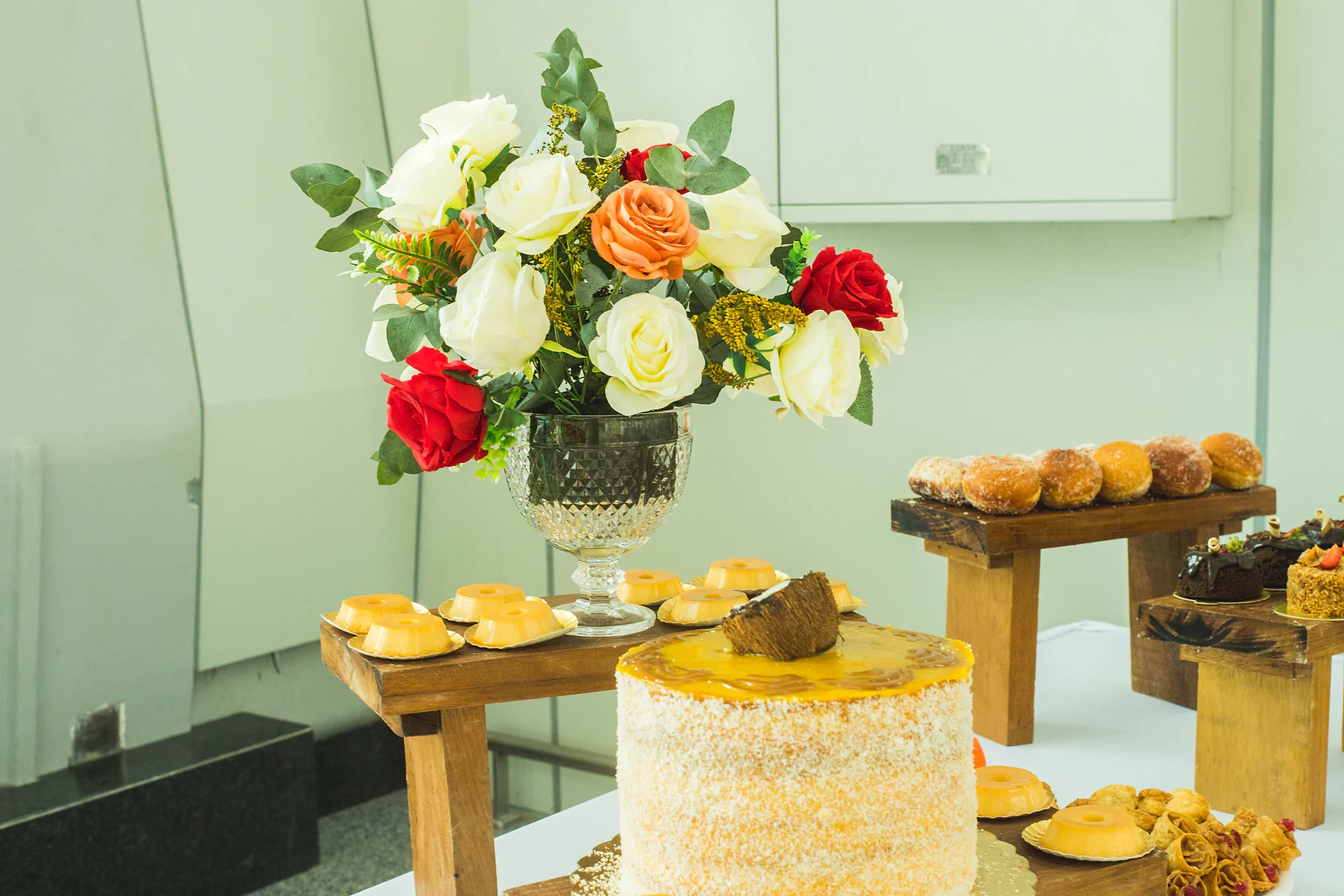 Arrangement of white, orange and red roses in a crystal goblet, beside a coconut cake and sweets on rustic wooden risers