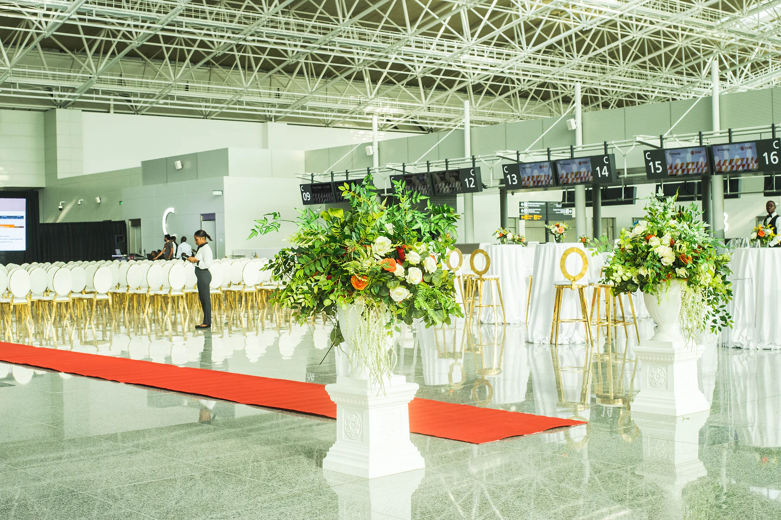 Two white pedestals with large arrangements of roses and foliage beside the red carpet, with tall tables and gold stools behind