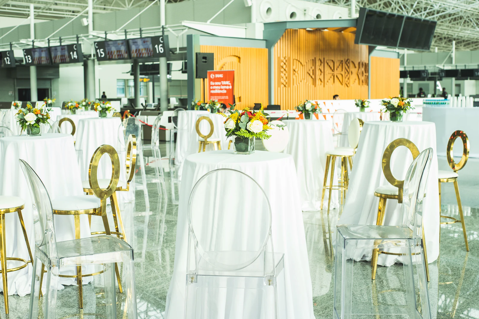 Tall tables in white cloths with small orange and white flower arrangements, clear chairs and gold-ringed stools in the hall