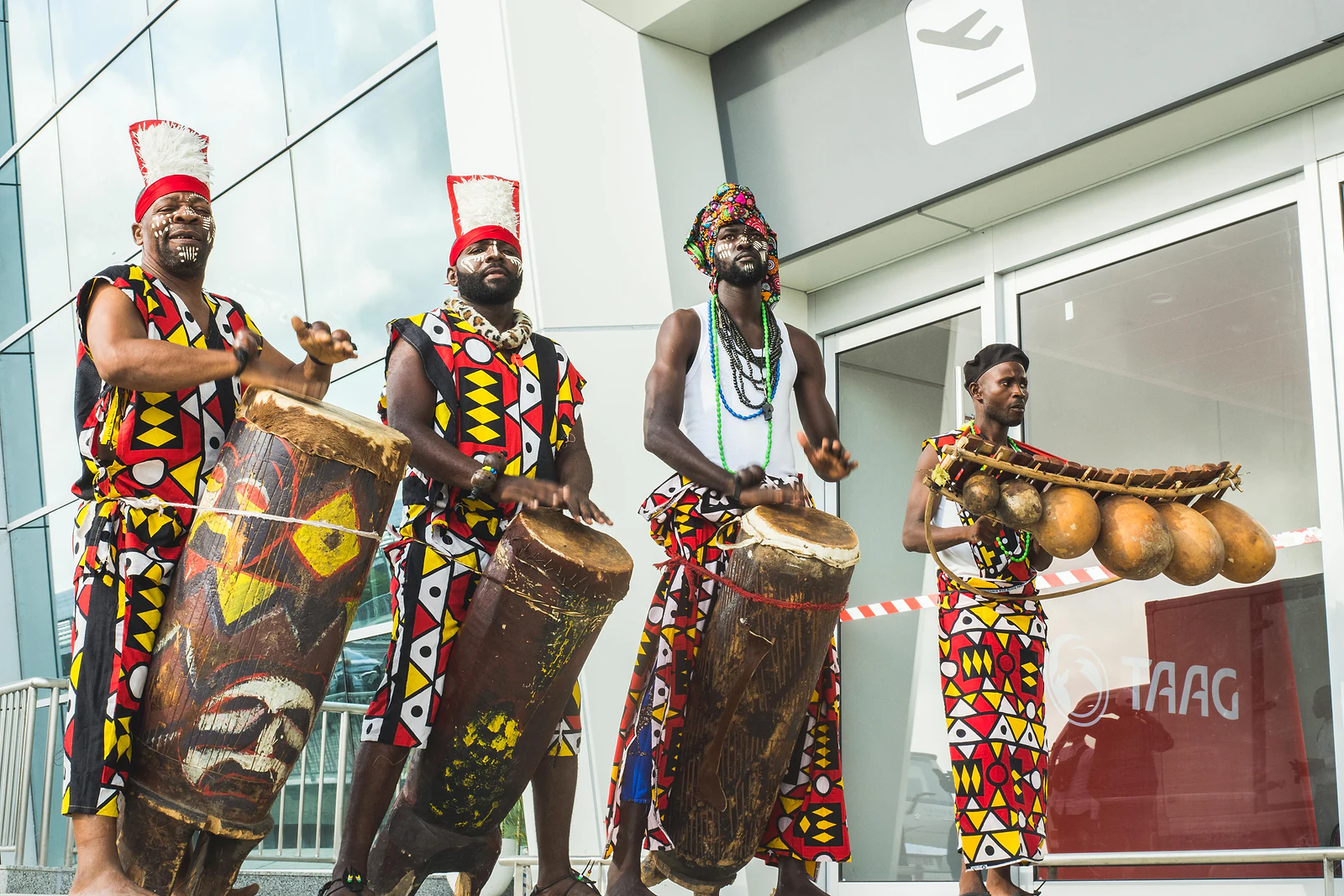 Four musicians in red and yellow geometric-print costumes, with painted drums and a gourd instrument, at the entrance