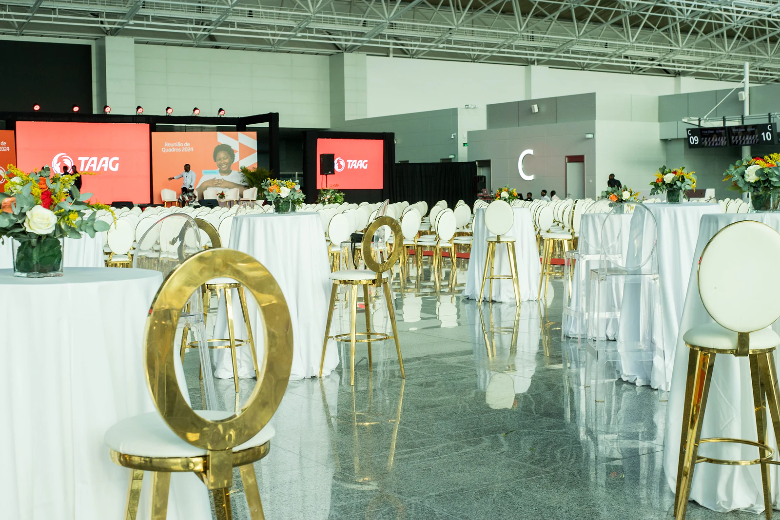 Gold-ringed stool and a tall white-clothed table with a flower arrangement, with the stage’s orange screens behind