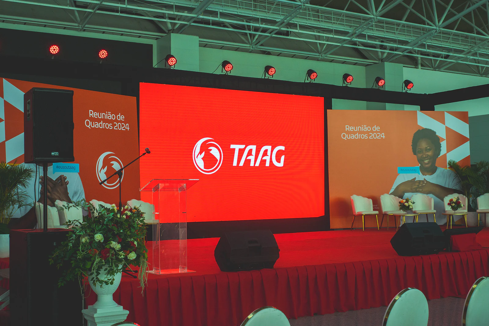 Stage with the large red TAAG screen, a clear lectern, a red stage skirt and an arrangement of white flowers in the foreground