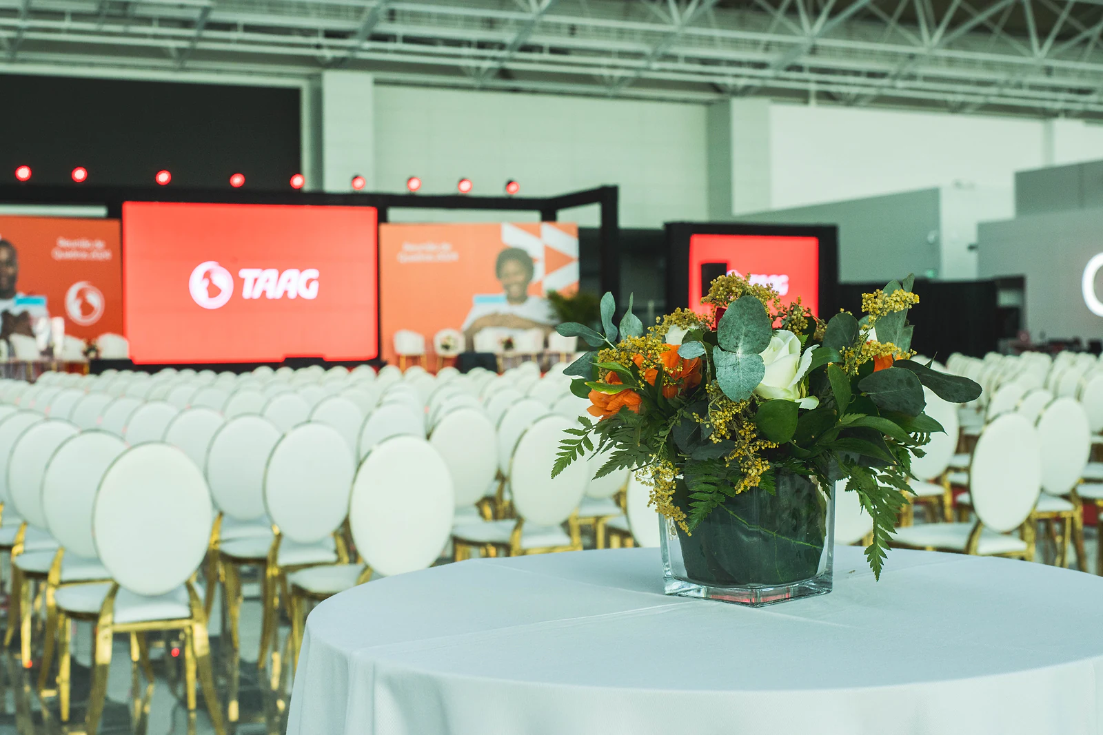 Arrangement of orange and white flowers with foliage on a white-clothed table, before the rows of chairs and the stage screens