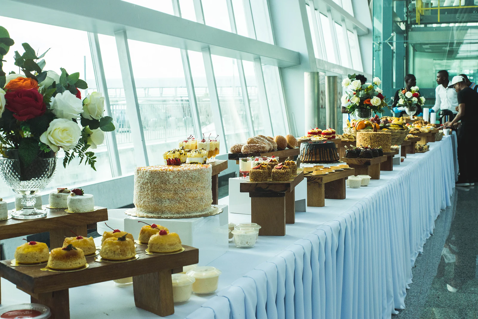 Long buffet table with a pleated white skirt, cakes, pastries and sweets on wooden risers and rose arrangements, beside the windows