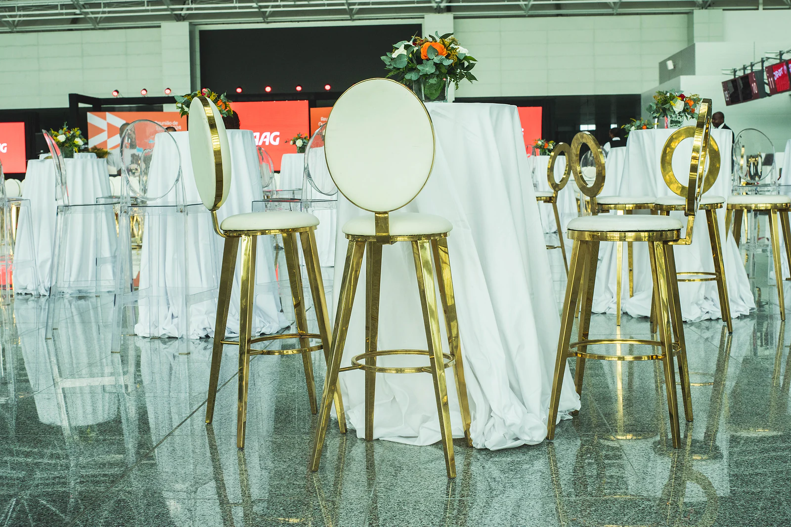 Tall gold stool with a round white back beside a tall white-clothed table with an orange flower arrangement, below the stage screens