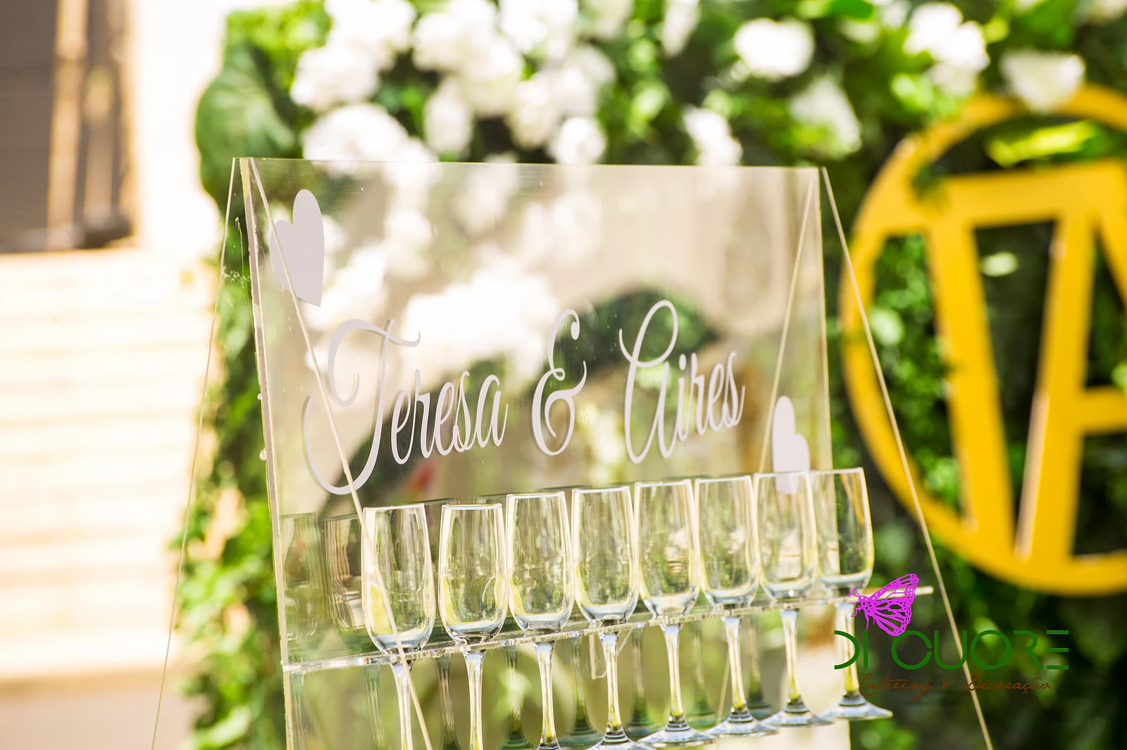 Acrylic sign bearing the names Teresa & Aires and a row of champagne flutes, in front of the foliage panel with the gold monogram
