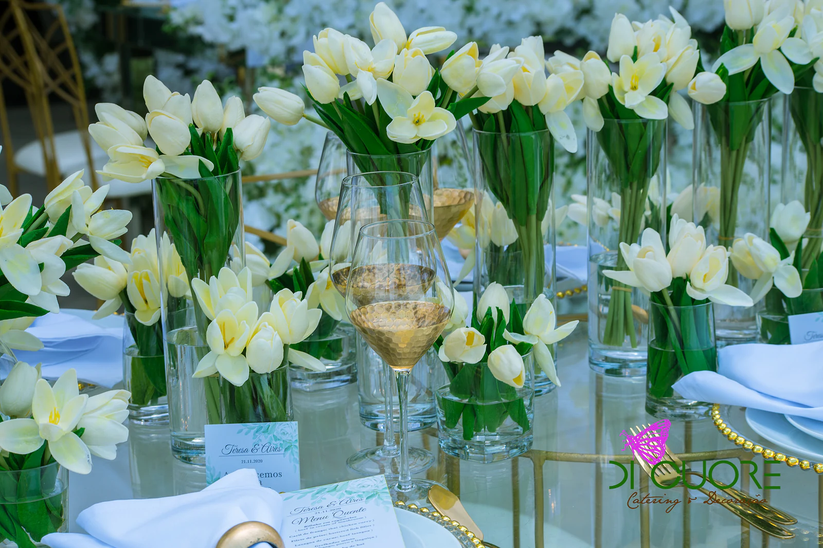 Tall vases of white and pale yellow tulips beside a golden glass, on a glass table, with a table card and menu