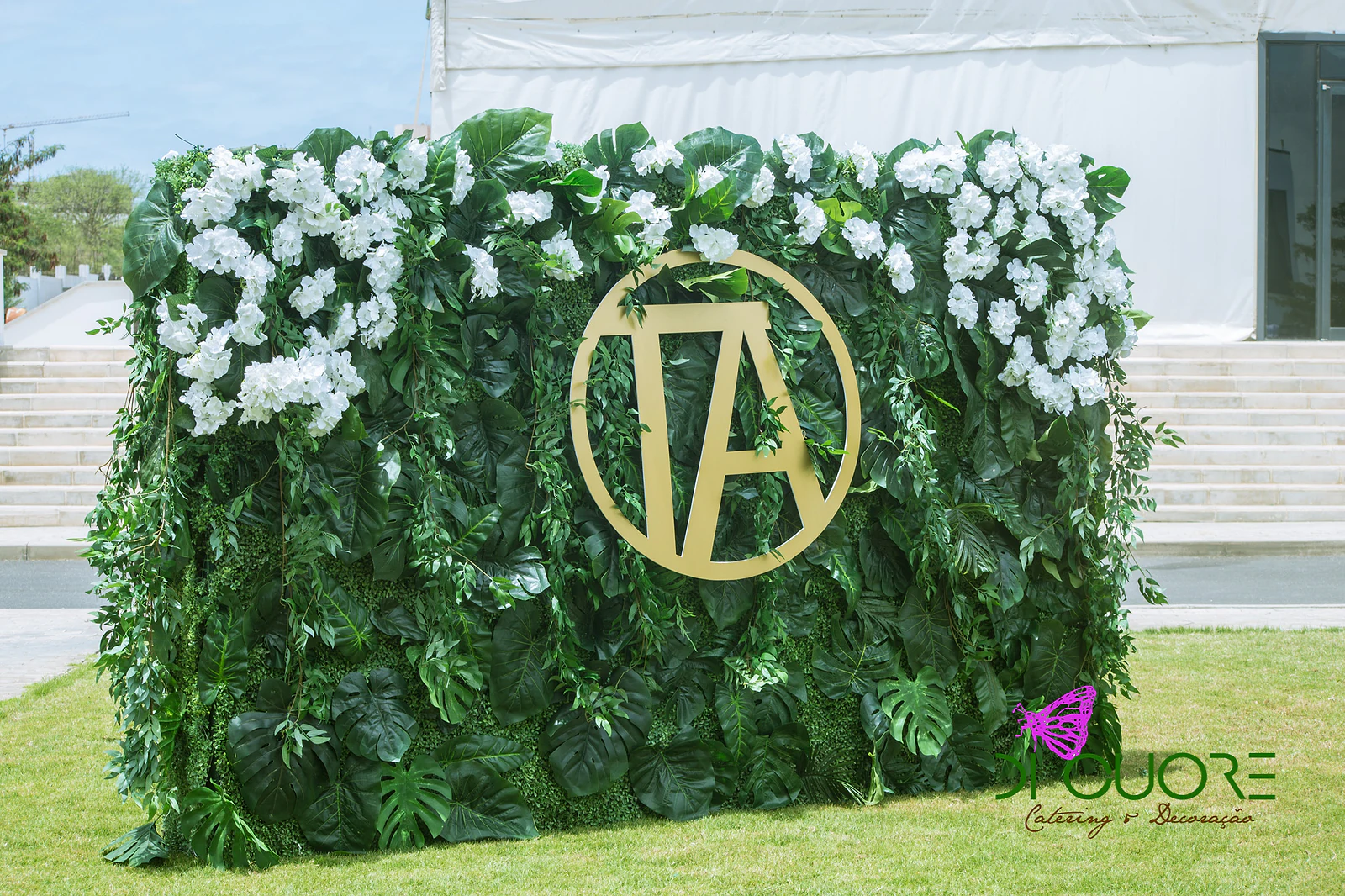 Green foliage panel with white flowers along the top and the gold TA monogram at its centre, standing on the lawn