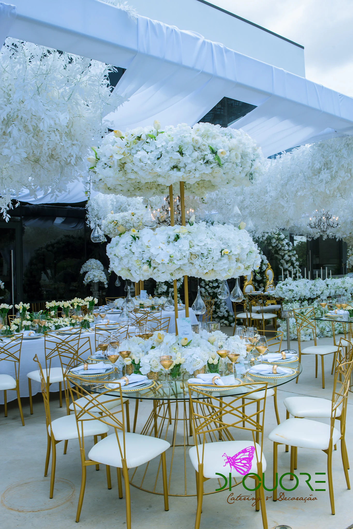 Round glass table with gold chairs with white seats, beneath a two-tier pillar of flowers and a ceiling of hanging white flowers