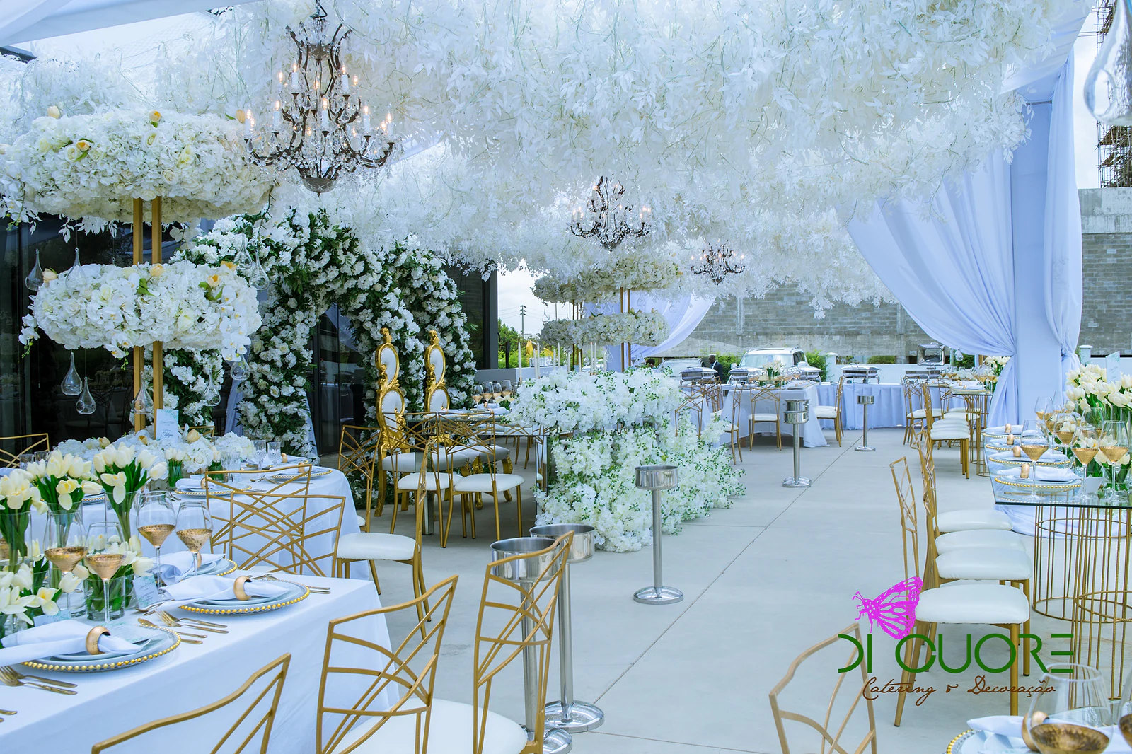 Aisle between laid tables with tulips and golden chairs, with two-tier pillars of flowers, chandeliers and flower arches at the far end