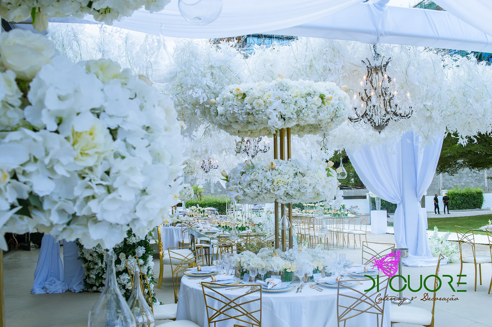 Large spray of white hydrangeas in the foreground and, beyond, two-tier pillars of flowers, a dark chandelier and round tables in white cloths
