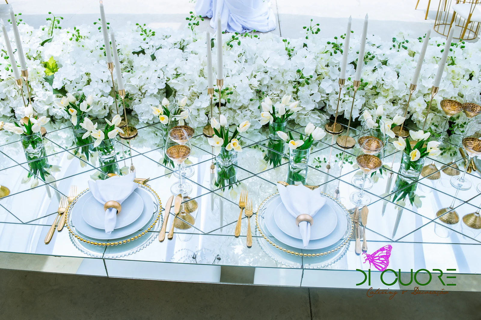 Mirror-topped table with plates, gold napkin rings, slim candles and small vases of tulips, beneath a band of white flowers