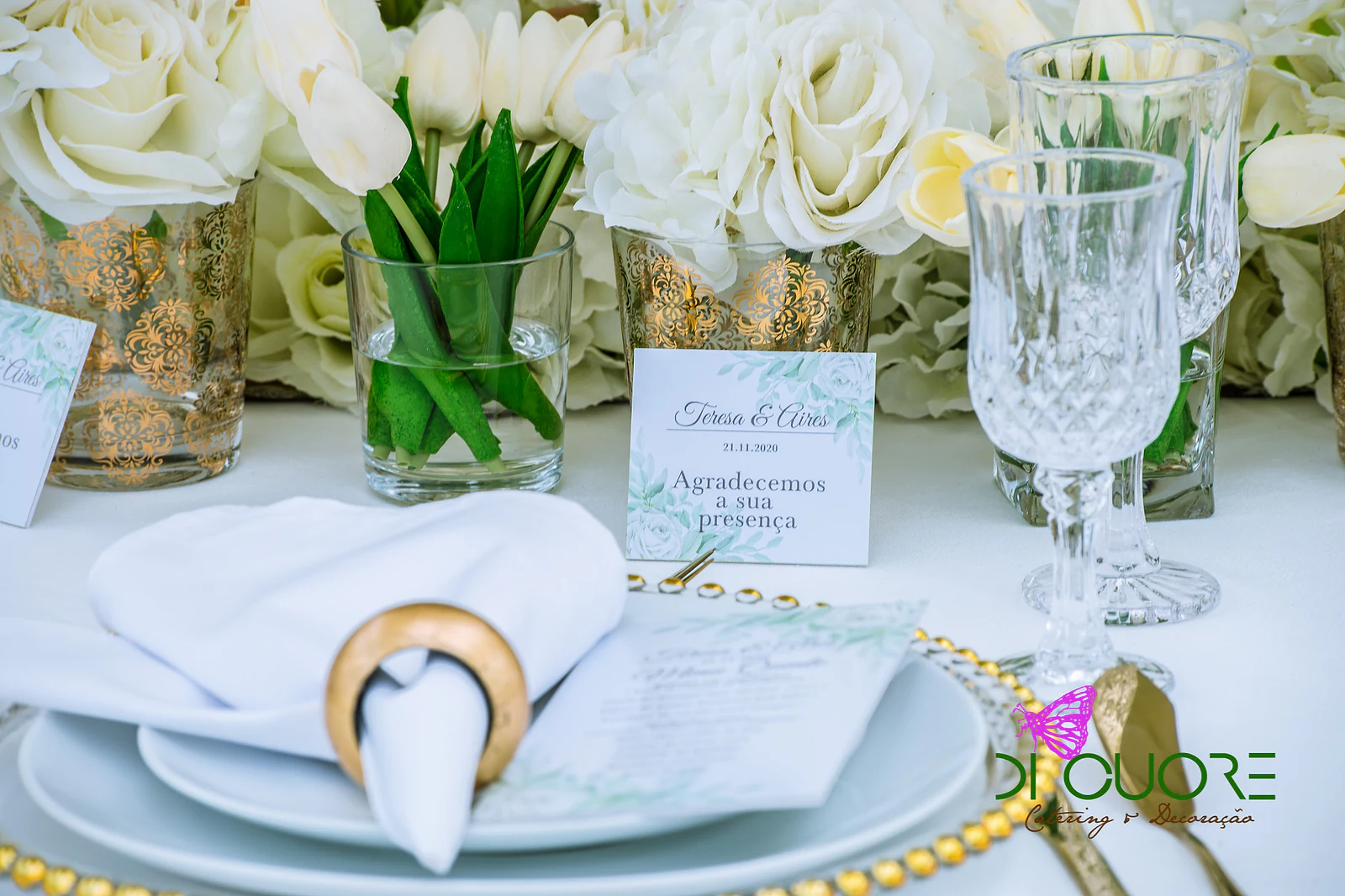 Plate on a gold beaded charger, white napkin in a gold ring and menu, beside vases of tulips and white roses and a thank-you card