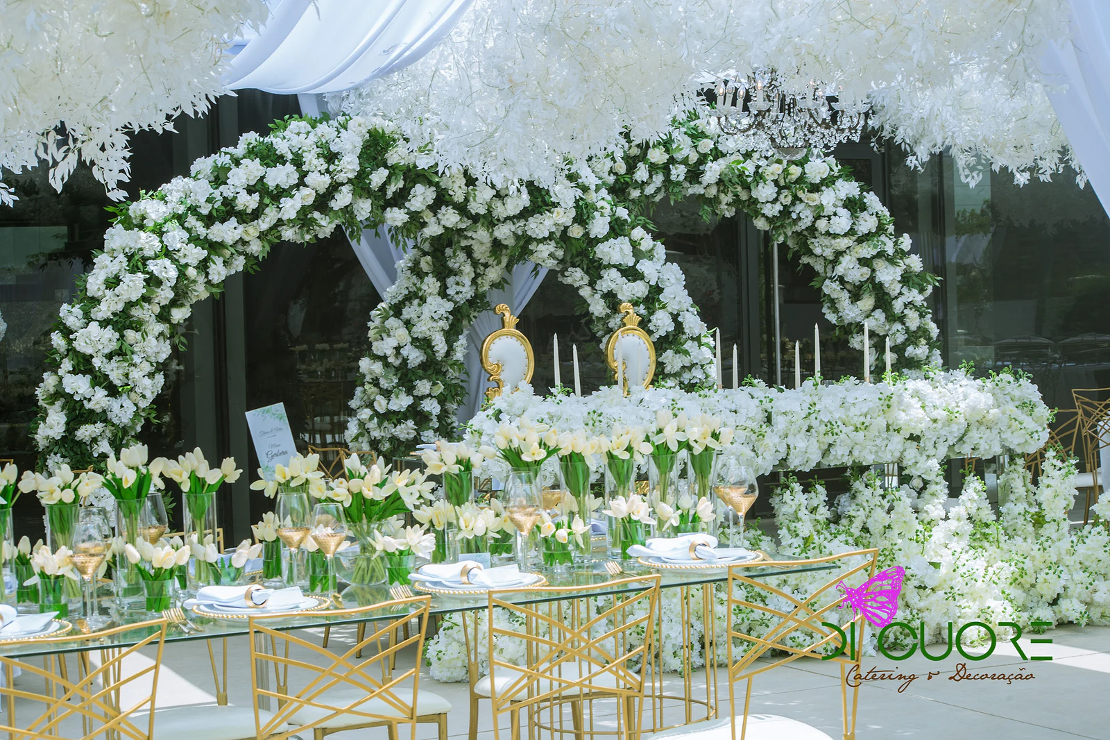 Two arches of white flowers and foliage above a table of white and gold armchairs, with tulips, candles and a band of white flowers in front, beneath a ceiling of hanging flowers