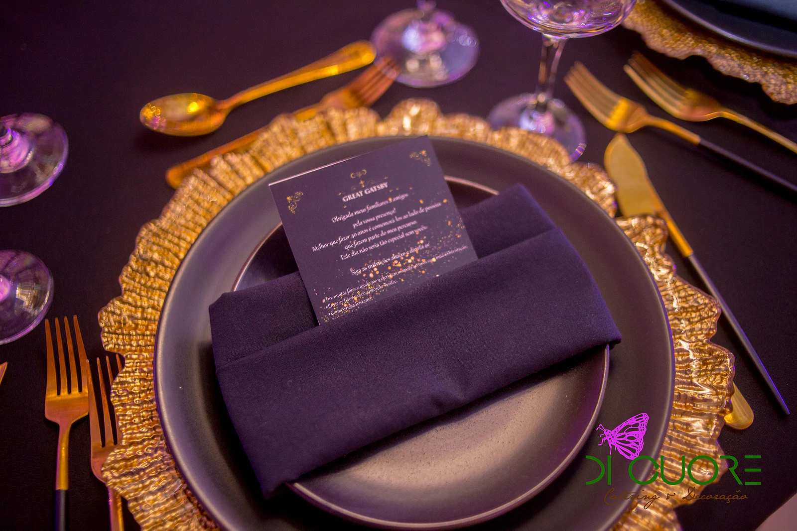 Prato preto sobre prato de presença dourado de rebordo ondulado, com guardanapo escuro, cartão de menu e talheres dourados sobre toalha preta