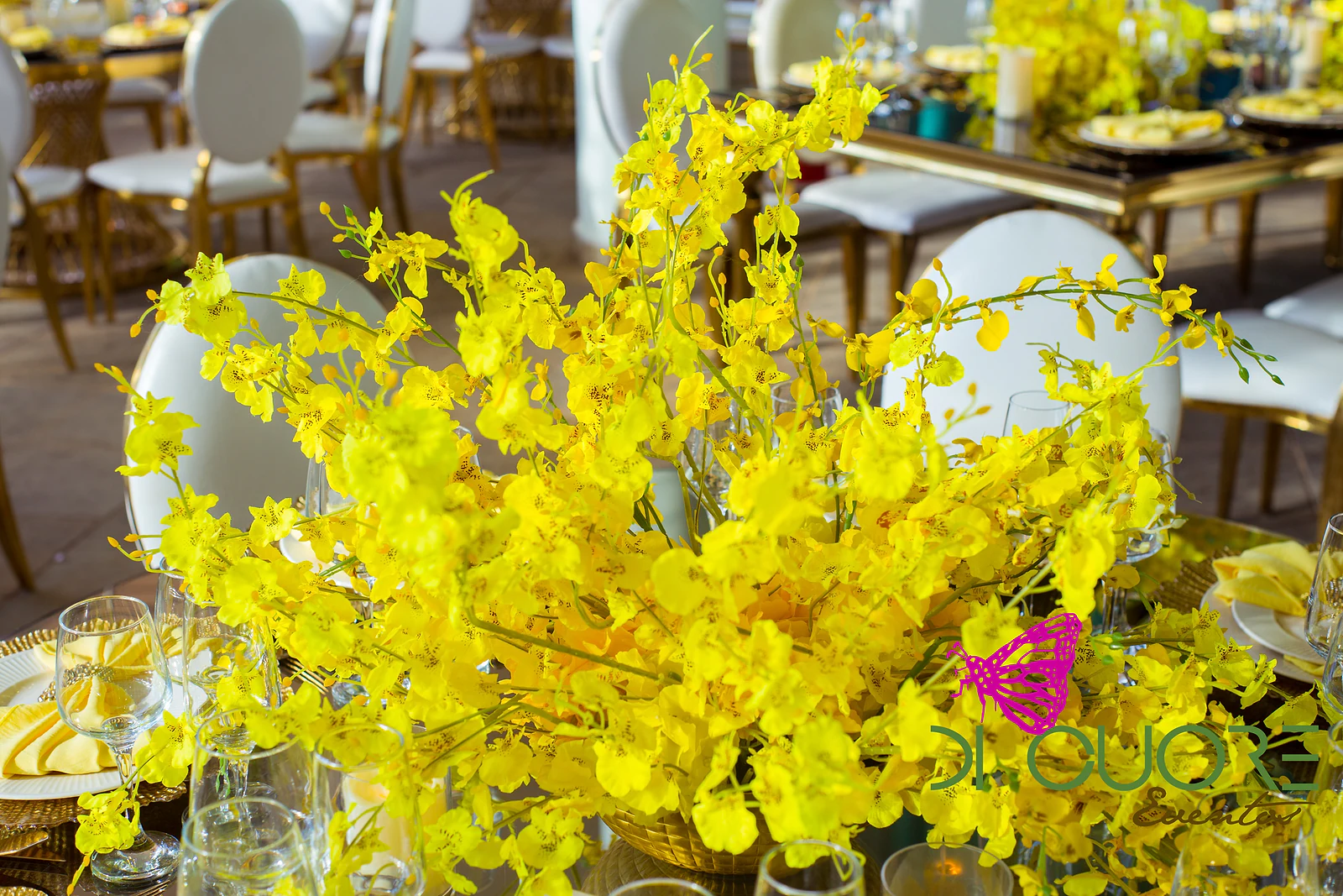 Dense spray of yellow orchids in the foreground, with gold-legged tables and round-backed chairs blurred behind