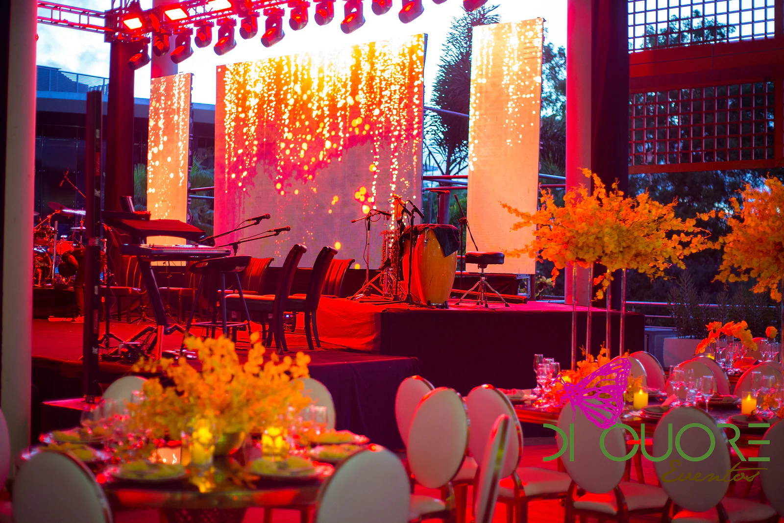 Open-air stage with panels of cascading orange and red lights and, in the foreground, tables with arrangements of yellow orchids at dusk