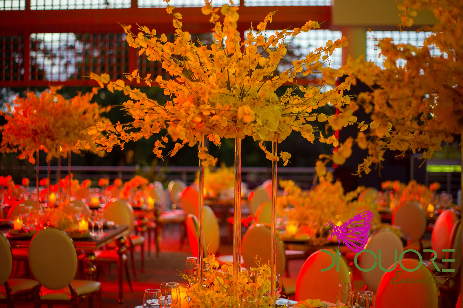Tall arrangements of yellow orchids on long tables, tinted orange by the terrace’s red lighting at dusk